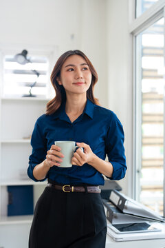 Smiling  asian businesswoman drink coffee enjoy morning in office. Happy female small business Entrepreneur look in distance thinking.