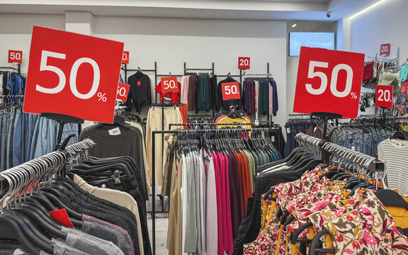 Large red 50 and 20 sale signs between racks of colorful women clothing, floral jackets, sweaters, jeans on hangers in fashion boutique, Black Friday, concept of discounts, retail promotion