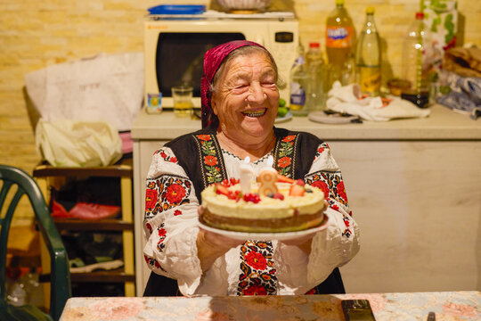 Portrait of an elderly Romanian woman in traditional costume holding a cake, celebrating her 81st birthday.