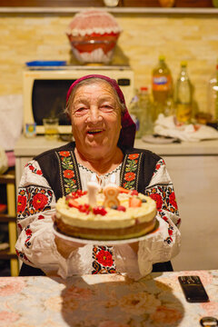 Portrait of an elderly Romanian woman in traditional costume holding a cake, celebrating her 81st birthday.