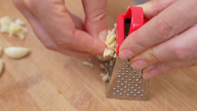 Close view of hands rubbing a peeled garlic clove on a small metal grater with a red handle, leaving fine shreds on a wooden kitchen board beside spare segments.