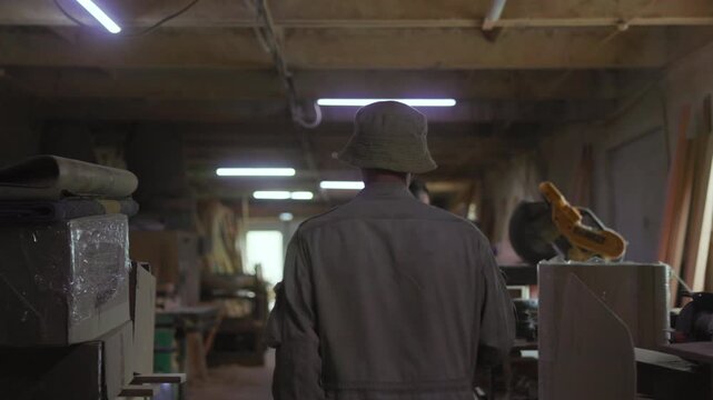 Rear view of person in hat and dark jacket standing in dim woodshop aisle under fluorescent lights, surveying workbenches and stacked wooden planks