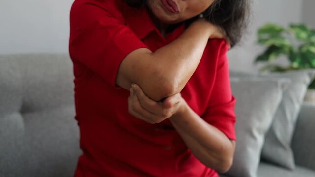 Close-up video of an Asian senior woman holding and massaging her painful elbow at home, illustrating elbow pain, joint discomfort, stiffness, and elderly healthcare.