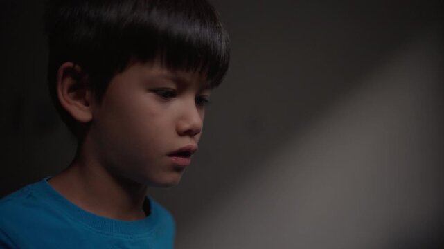 A boy in a blue t-shirt looks down quietly, then lifts his gaze upward before lowering his eyes again against a dark wall