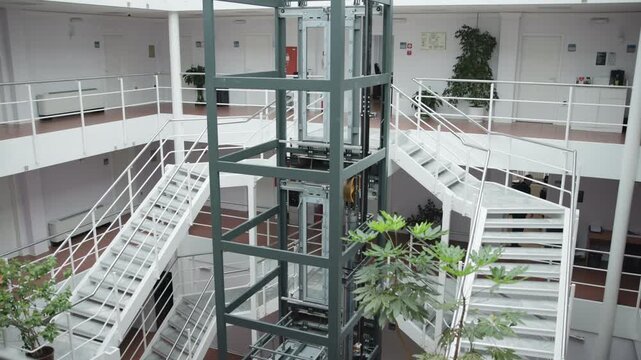 Exposed elevator shaft and white metal staircase spans across multi level atrium with potted plants under cool daylight