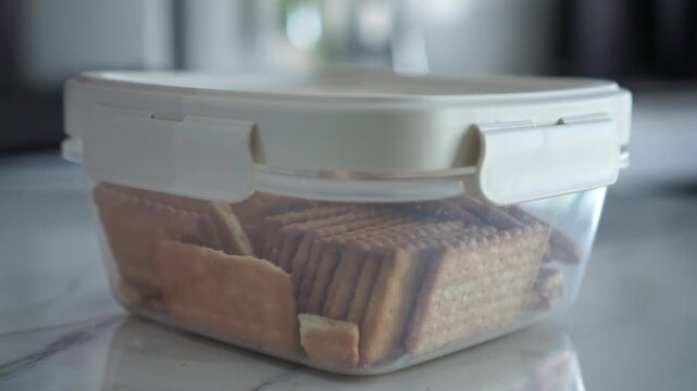 Hand unlocking a clear storage box with biscuits and crackers inside. Household footage capturing food organization, pantry, and daily routine.