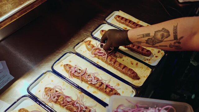 Rows of grilled kebabs on lavash bread being garnished with red onion under the warm light of a kitchen pass.