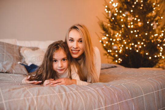 Happy woman with child girl in bed over Christmas tree with lights in room. Winter holiday season.