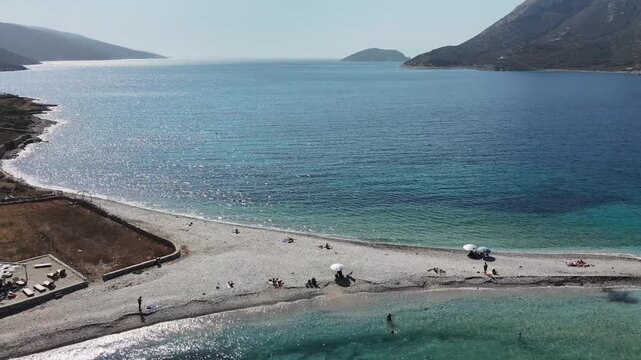 Aerial drone shot of the narrow gravel spit of Agios Pavlos beach reaching into the clear turquoise sea with people bathing, off the north coast of Amorgos, Cyclades, Greece