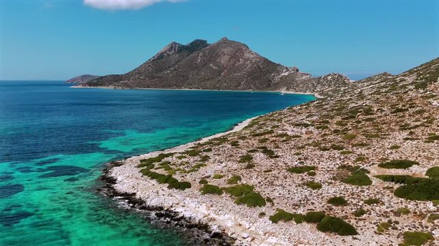 Aerial drone shot of the uninhabited islet of Nikouria across a narrow calm strait of clear turquoise water from the rocky shore of Agios Pavlos on Amorgos, Cyclades, Greece