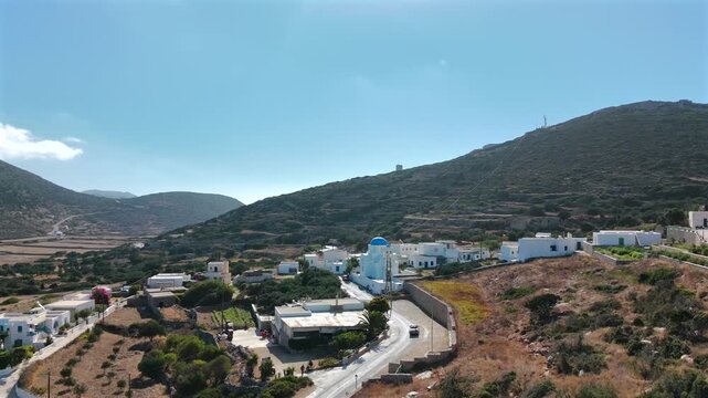 Aerial drone shot flying toward the village of Arkesini with the blue-domed church of Agios Onoufrios and a car on the road, Kato Meria, southern Amorgos, Cyclades, Greece