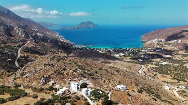 Aerial drone shot flying over an arid terraced hillside toward the deep Aegiali bay with turquoise shallows and the mountainous islet of Nikouria on the north coast of Amorgos, Cyclades, Greece
