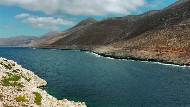 Aerial drone shot of the strait between Amorgos and Nikouria islet with a coastal road on the steep rugged mountainside and the distant bay of Aegiali, Cyclades, Greece, on a sunny day