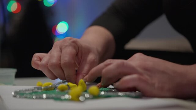 A woman's hands thread silver tinsel between yellow pom poms on a green glitter star craft as Christmas lights glow in the background, Low Angle
