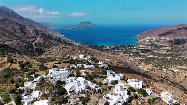Aerial drone shot flying past the church of Agioi Anargyroi over the white cubic houses of Tholaria toward Aegiali bay and Nikouria islet on Amorgos, Cyclades, Greece