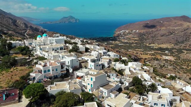 Aerial drone shot flying toward the whitewashed village of Tholaria with the blue-domed church of Agioi Anargyroi above Aegiali bay and Nikouria islet on Amorgos, Cyclades, Greece