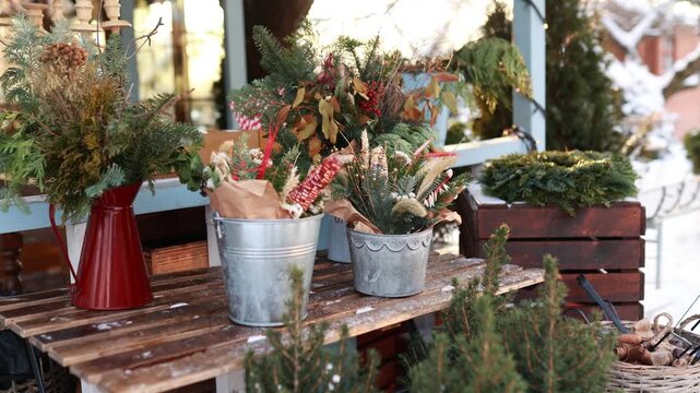 Cozy Сhristmas country house porch featuring evergreen floral bouquets in red jug and zinc buckets on wooden bench. Festive xmas holiday terrace decor concept, snowy background, copy space.