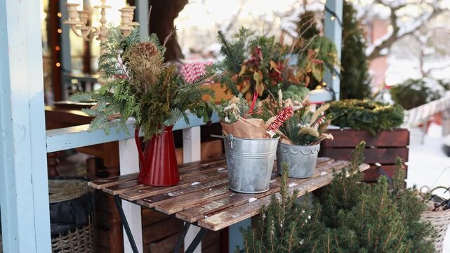 Cozy Сhristmas country house porch featuring evergreen floral bouquets in red jug and zinc buckets on wooden bench. Festive xmas holiday terrace decor concept, snowy background, copy space.   