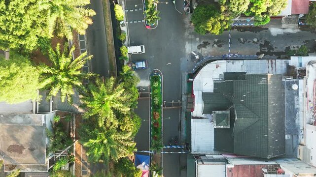 Aerial view of a quiet residential street in Malang, East Java, Indonesia, featuring lush palm trees, rooftops, and a narrow road with a central green median.