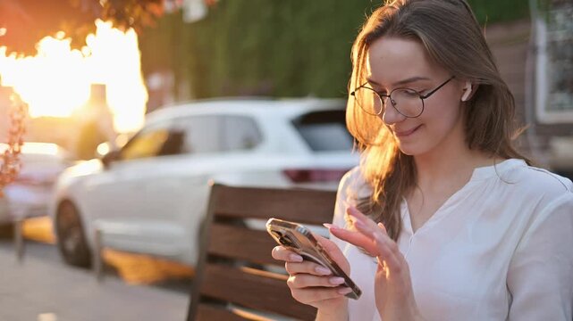 Young Caucasian woman with glasses sitting on a bench outdoors, holding a smartphone, with sunlight filtering through trees and cars parked in the background