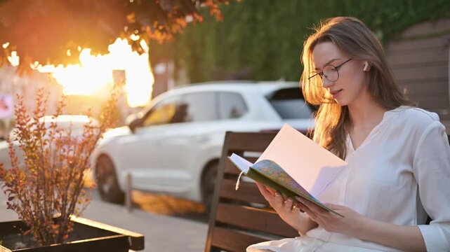 A young woman sits on a wooden bench reading a small book at sunset, wearing glasses and wireless earbuds. Warm golden-hour light and an urban background create a peaceful, contemplative mood.