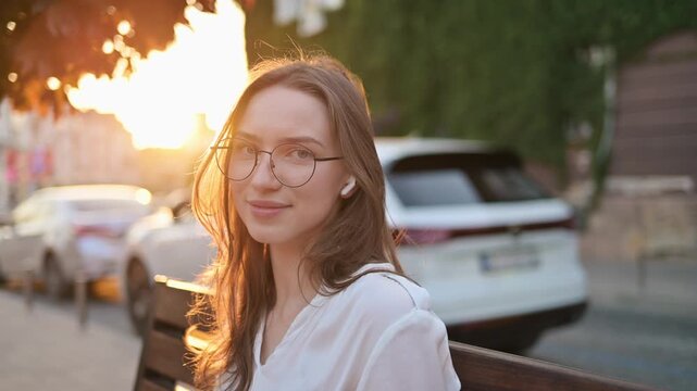 Young woman with glasses relaxes on a wooden bench in a city park, with flowering plants and parked cars visible in the background during sunset