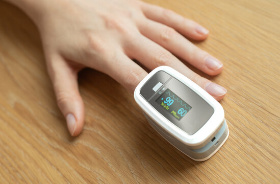 Pulse oximeter sensor clipped to fingertip reads oxygen saturation at 99% and pulse rate 60. Portable SpO2 device monitors vital signs as hand rests on wooden table. Compact gadget for home checkup.
