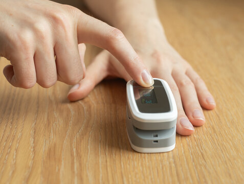 Index finger presses button on pulse oximeter measuring oxygen saturation and heart rate. Fingertip clip sensor tracks vital signs as hand steadies device on wooden. Portable gadget, home checkup.
