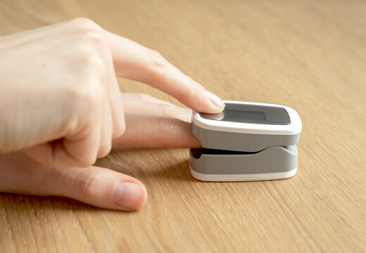 Finger pressed into pulse oximeter clip measures blood oxygen saturation level. Fingertip sensor checks heart rate and vital signs on a wooden table. Portable digital device for home health checkup.