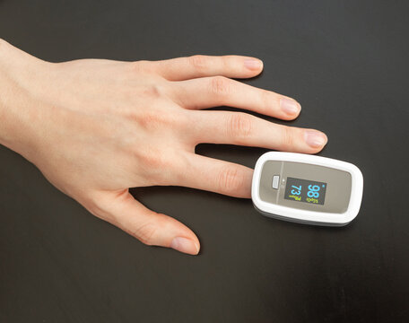 Oxygen saturation and pulse rate glow on a fingertip oximeter sensor. Hand rests on dark surface as SpO2 monitor tracks vital signs. Top view, compact digital device for self test.