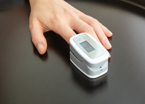 Pulse oximeter clip on finger displays oxygen saturation and heart rate readings. SpO2 sensor monitors vital signs with hand resting on dark glossy table. Compact digital device for home checkup.