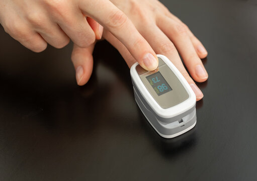 Fingertip pulse oximeter reads oxygen saturation 98 and heart rate 73. Outstretched hand reaches onto sensor device, SpO2 monitor glowing on dark glossy surface. Close up top view, home checkup.