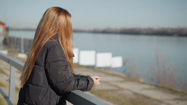 Woman with long hair stands by railing gazing out over calm water in outdoor setting. Woman wears dark puffer jacket and maintains same pose throughout hands clasped on rail against soft