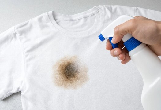 A person spraying a chemical stain remover on a stubborn dark stain on a white cotton t-shirt during laundry preparation