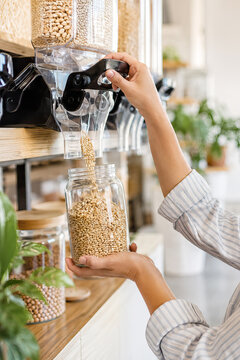 A woman carefully dispenses grains from a clear plastic unit into a glass jar, holding the jar with her hand within a bright, modern kitchen. zero footprint, eco-friendly