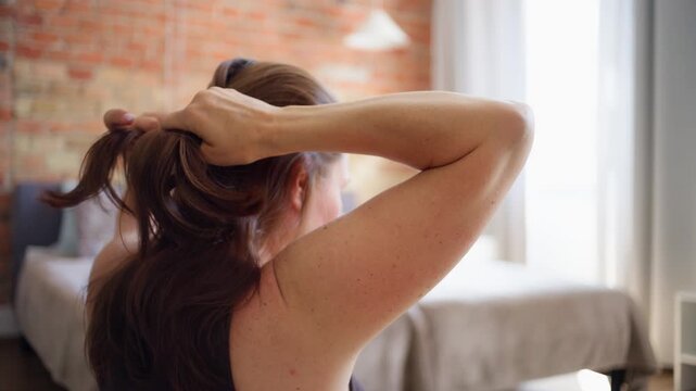 Young woman gathering her long brown hair to tie a ponytail in a sunny bedroom during daytime