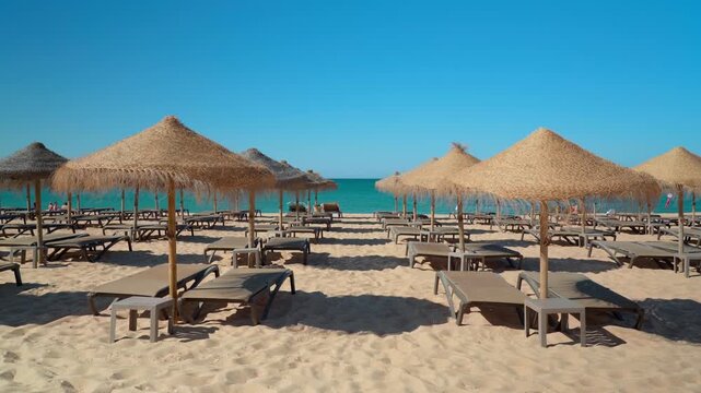 Rows of empty sunbeds and straw umbrellas on a sandy beach waiting for tourists on a sunny day. Beautiful turquoise sea and clear blue sky in a famous european summer vacation destination