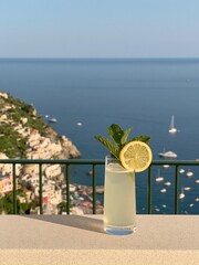 Fototapeta premium Refreshing Lemonade Cocktail with Mint Garnish on Balcony Overlooking Italian Coastline and Ocean