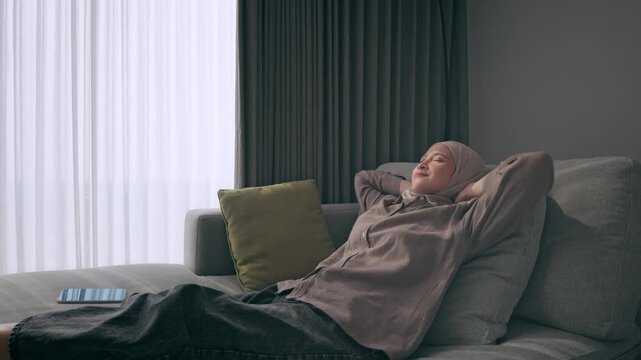 Asian Muslim woman wearing a hijab leans back on a comfortable sofa in a modern living room, eyes closed and smiling as she enjoys a quiet moment of relaxation and calm at home.