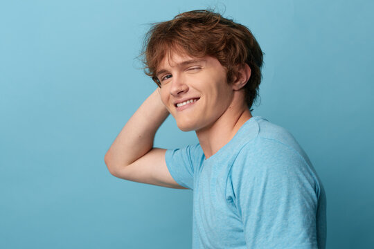Bashful young man with wavy brown hair grinning and scratching head behind neck, wearing a blue t-shirt on a light blue background. Concept of modest embarrassment and flattered reaction.