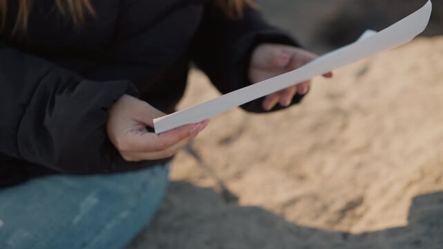 Woman examines blank sheet of paper while seated outdoors on sand. Woman holds white document with both hands focusing intently as woman inspects its contents against natural daylight