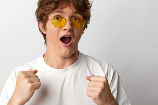 Astounded young man in yellow tinted sunglasses and white t-shirt, mouth open in shock, fists clenched near chest against plain grey background, emotional reaction