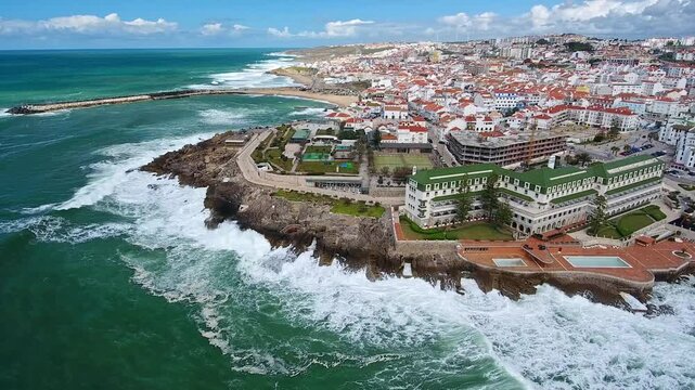 Aerial drone view flying over the scenic coastal town of ericeira in portugal, with powerful atlantic ocean waves breaking against the rocky coastline and beautiful architecture