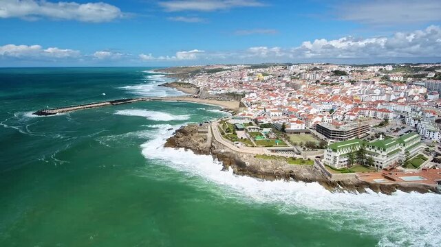 Dramatic aerial view of the charming seaside town of ericeira, portugal, with powerful atlantic ocean waves crashing against the rugged cliffs and the beautiful baleia beach on a sunny day