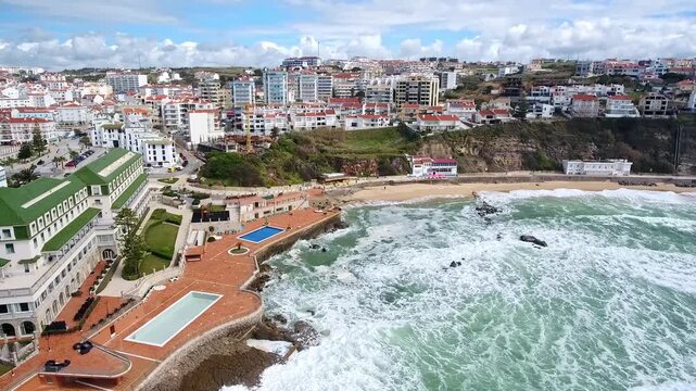 Beautiful panoramic aerial shot flying over the coastal town of ericeira in portugal, showing strong atlantic waves crashing on the cliffs and the famous baleia beach under a cloudy sky