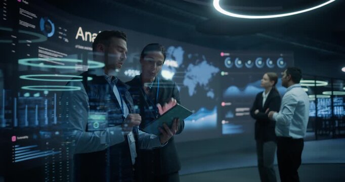 White and Asian Analysts Review a Tablet While and Discuss Sales Forecasts Beside Global Data Walls, Coordinating Market Expansion in a Modern Business Intelligence Command Center.