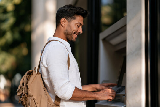 Businessman with short dark hair operates an ATM outdoors. Personal finance, card banking, cash withdrawal, professional lifestyle.