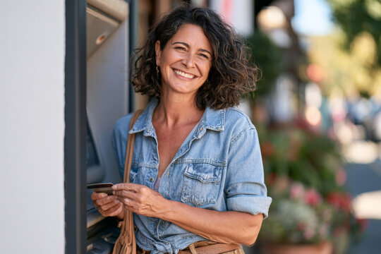 Smiling woman with curly dark hair holds a card beside an outdoor ATM. Personal banking, card payment, financial confidence, urban lifestyle.