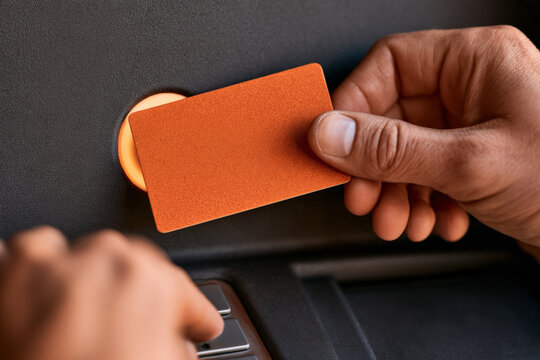 Hand presents an orange bank card beside an ATM card reader. Personal banking, card transaction, cash access, financial technology.