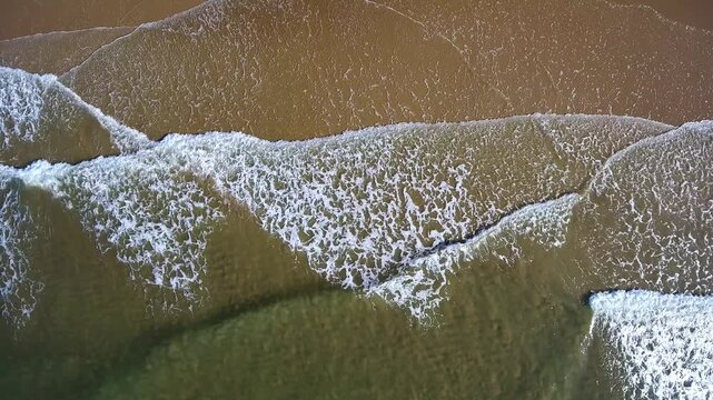 Calming aerial top down view of gentle ocean waves with white foam washing over a beautiful sandy beach, creating a relaxing and mesmerizing natural pattern on the shoreline during a sunny day